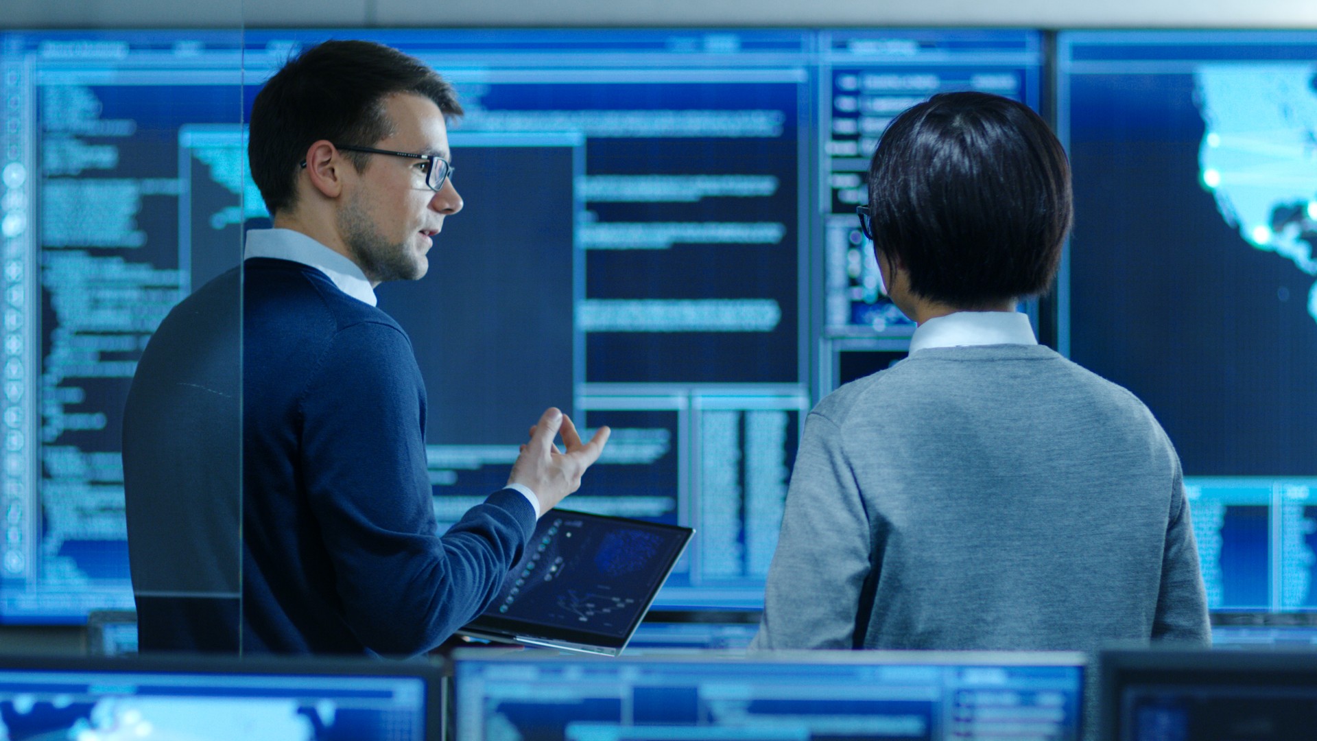 In the System Control Room IT Specialist and Project Engineer Have Discussion while Holding Laptop, they're surrounded by Multiple Monitors with Graphics. They Work in a Data Center on Data Mining, AI and Neural Networking.